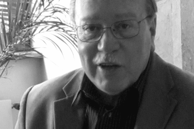 Portr&auml;t eines &auml;lteren Mannes mit Brille, der in einem Raum mit Pflanzen im Hintergrund spricht. Das Bild ist schwarz-wei&szlig;.