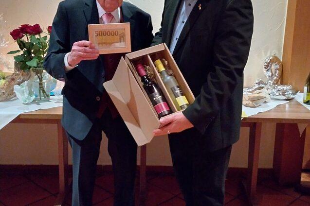 Zwei M&auml;nner stehen nebeneinander, einer h&auml;lt einen gro&szlig;en Scheck, der andere eine Geschenkverpackung mit Weinflaschen.