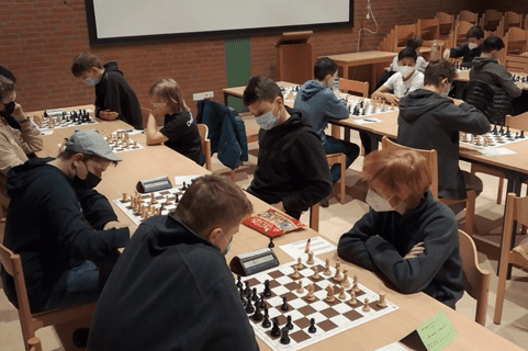 Schachturnier mit Jugendlichen, die konzentriert am Tisch spielen, umgeben von Schachbrettern und St&uuml;hlen.