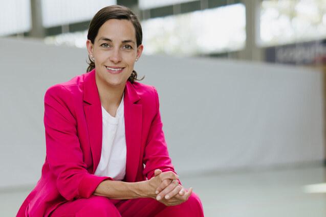 Frau in pinkem Anzug sitzt auf einer Bank, umgeben von Sportb&auml;llen: Basketball, Fu&szlig;- und Volleyball.