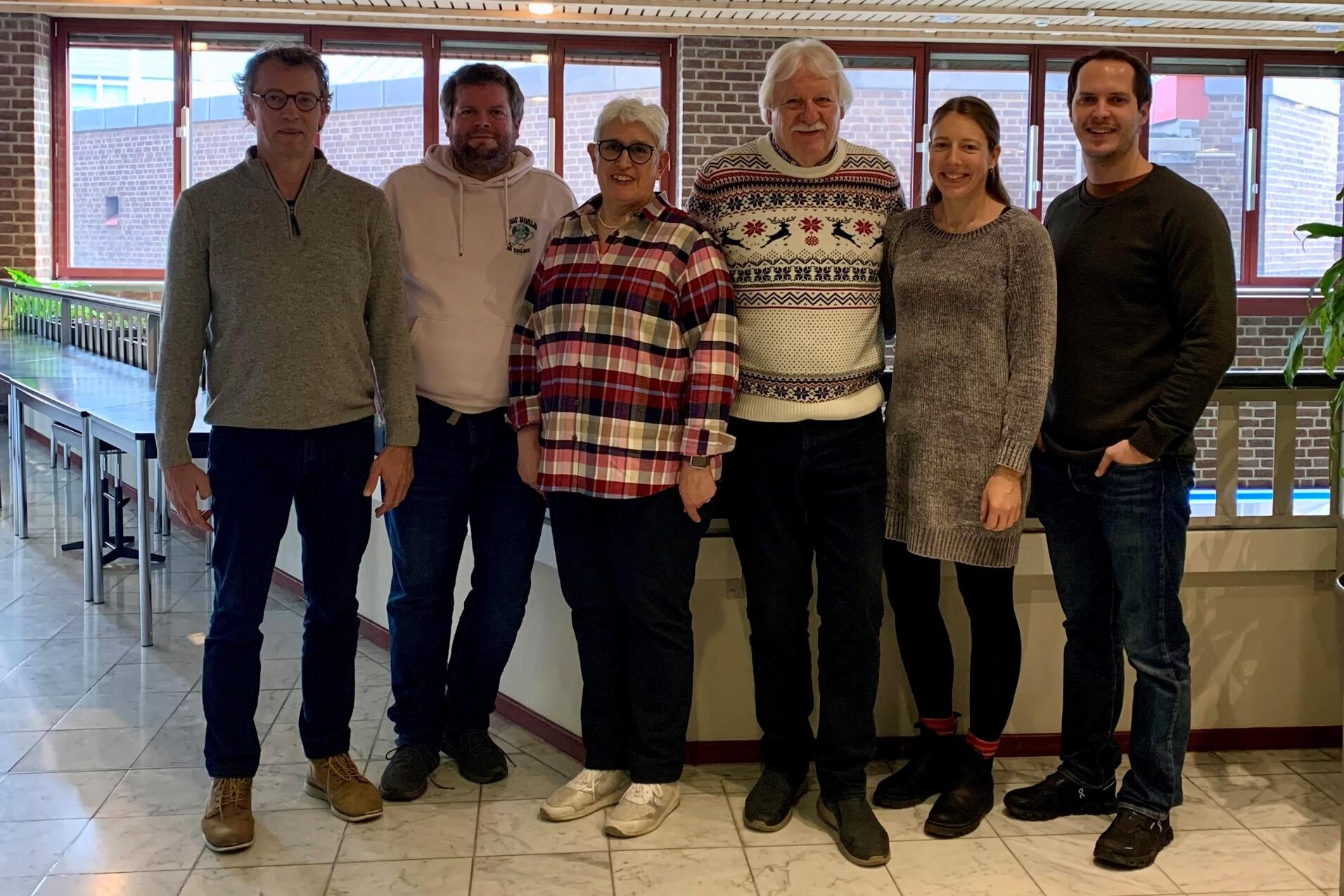 Gruppe von sechs Personen steht in einem modernen Innenraum mit Holzdecken und gro&szlig;en Fenstern.