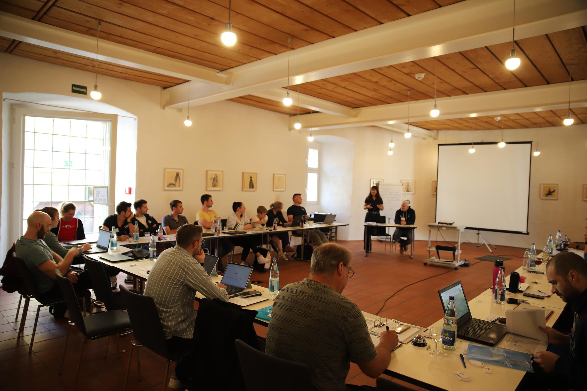 Gruppenarbeit in einem Seminarraum mit Teilnehmern an Tischen, Pr&auml;sentation an der Wand und Moderatorin vorne.