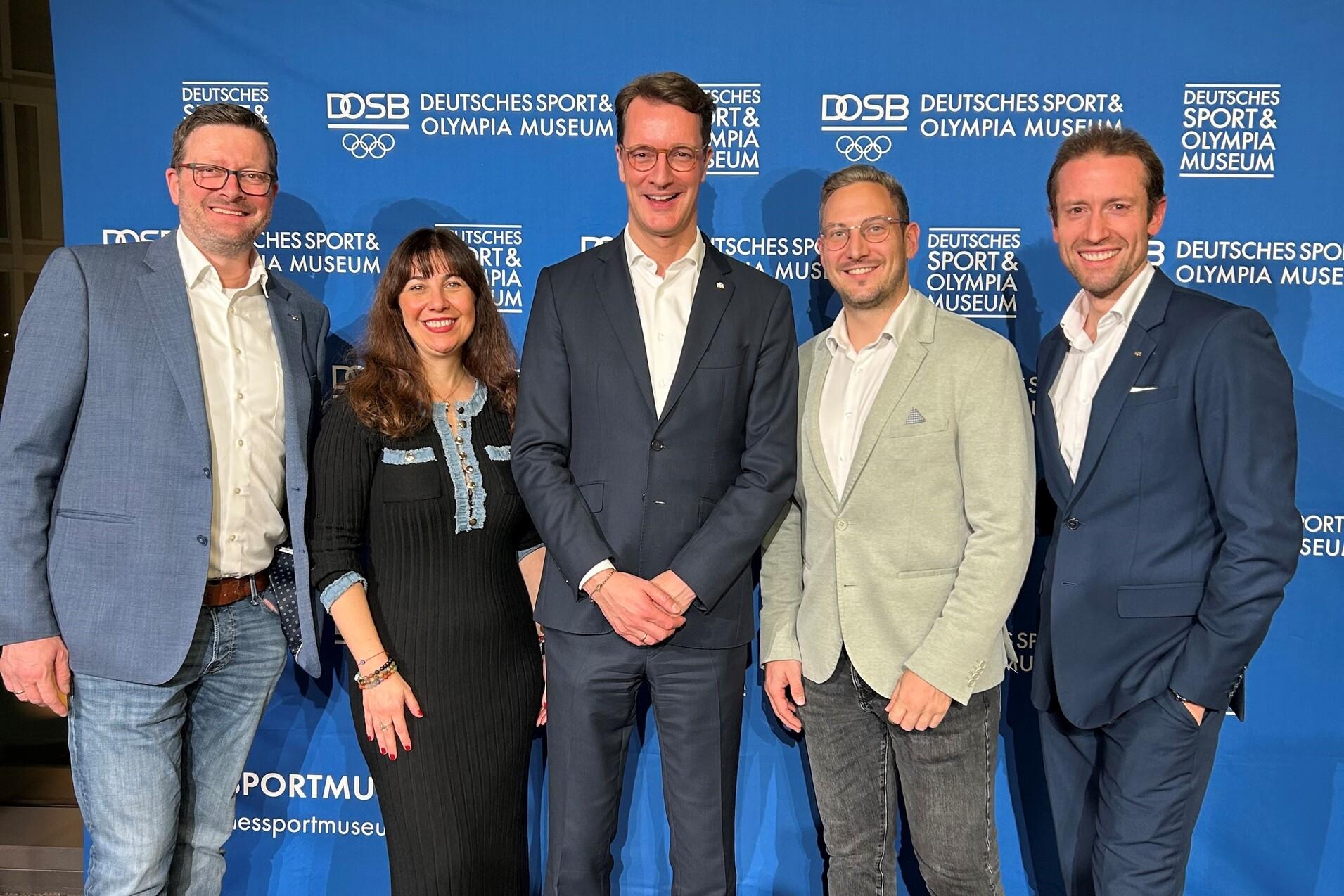 F&uuml;nf Personen posieren vor einer blauen Wand mit dem Logo des Deutschen Sport- und Olympiamuseums.