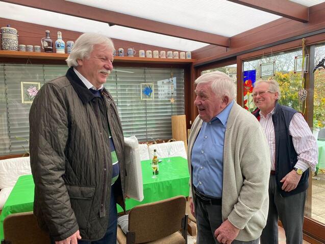 Drei &auml;ltere M&auml;nner unterhalten sich l&auml;chelnd in einem lichtdurchfluteten Wintergarten. Gr&uuml;ne Tischdecke im Hintergrund.