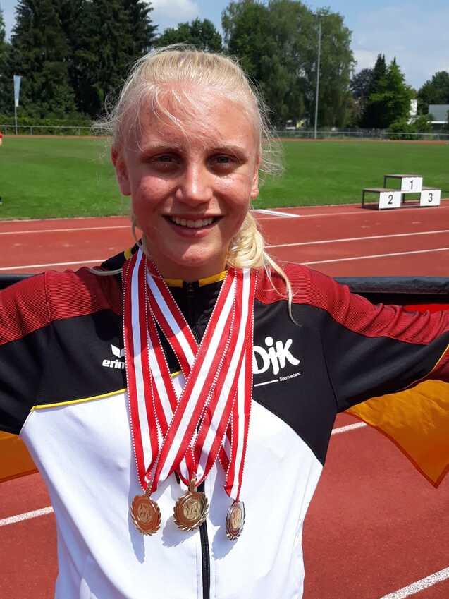 Jugendliche Athletin mit mehreren Medaillen um den Hals l&auml;chelt vor einem Sportfeld mit Laufbahn.