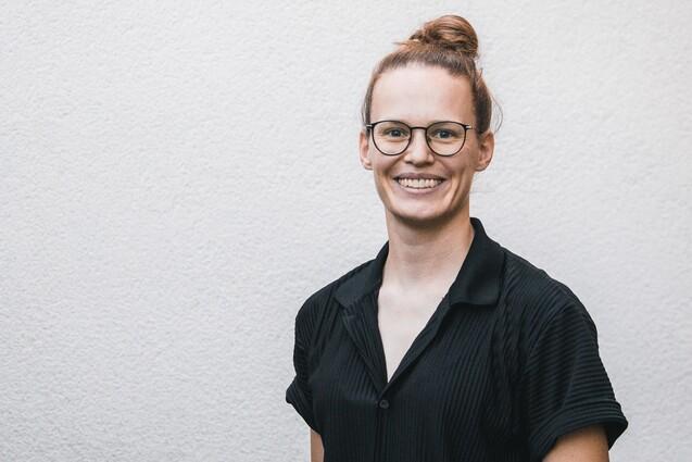 Frau mit schulterlangen, braunen Haaren im Dutt, tr&auml;gt eine schwarze Bluse und Brille, steht vor einer hellen Wand.