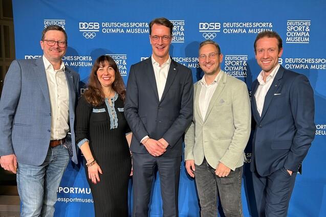 F&uuml;nf Personen posieren vor einer blauen Wand mit dem Logo des Deutschen Sport- und Olympiamuseums.