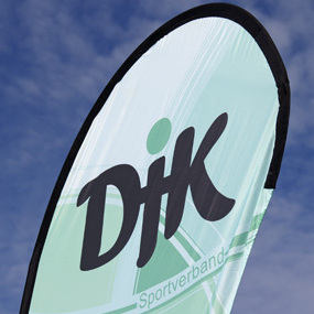 Fahne des DJK Sportverbands mit gr&uuml;nem Hintergrund und schwarzem Logo vor einem blauen Himmel.