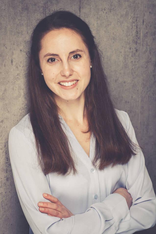 Junge Frau mit langen, braunen Haaren, l&auml;chelt in einem hellblauen Hemd vor einer grauen Betonwand.