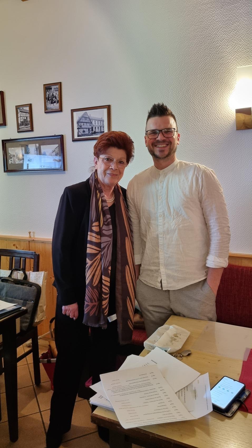 Eine Frau mit Brille und ein Mann mit Brille stehen l&auml;chelnd in einem Restaurant, umgeben von Bildern an der Wand.