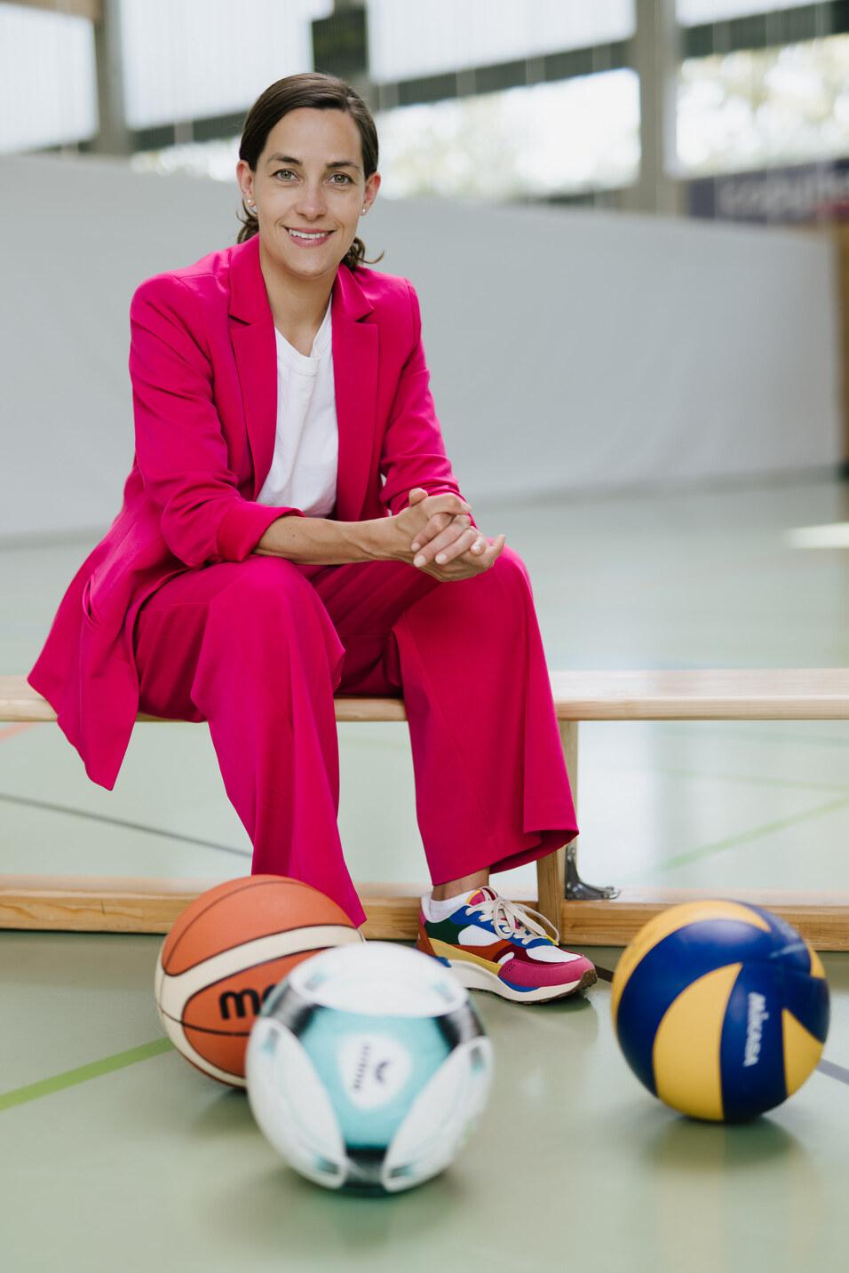 Frau in pinkem Anzug sitzt auf einer Bank, umgeben von Sportb&auml;llen: Basketball, Fu&szlig;- und Volleyball.