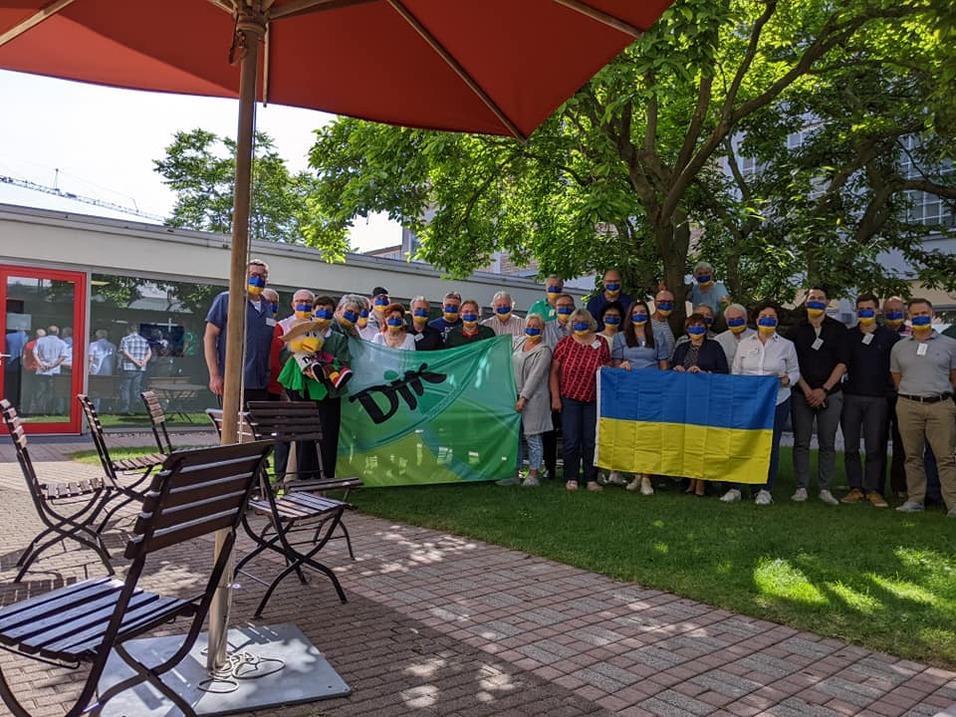 Gruppenfoto von Personen mit blauen und gelben Masken, die Fahnen halten, in einem Garten unter einem Sonnenschirm.