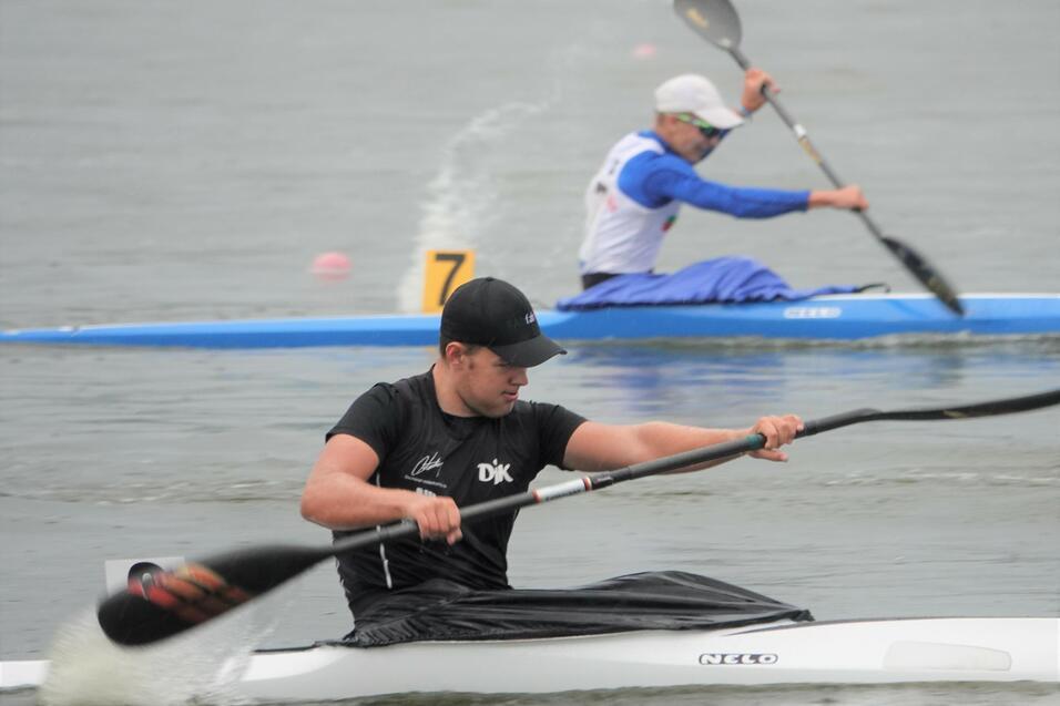 Zwei Kanufahrer paddeln auf einem Gew&auml;sser; einer in einem wei&szlig;en und blauen Kajak, der andere in einem schwarzen Kajak.
