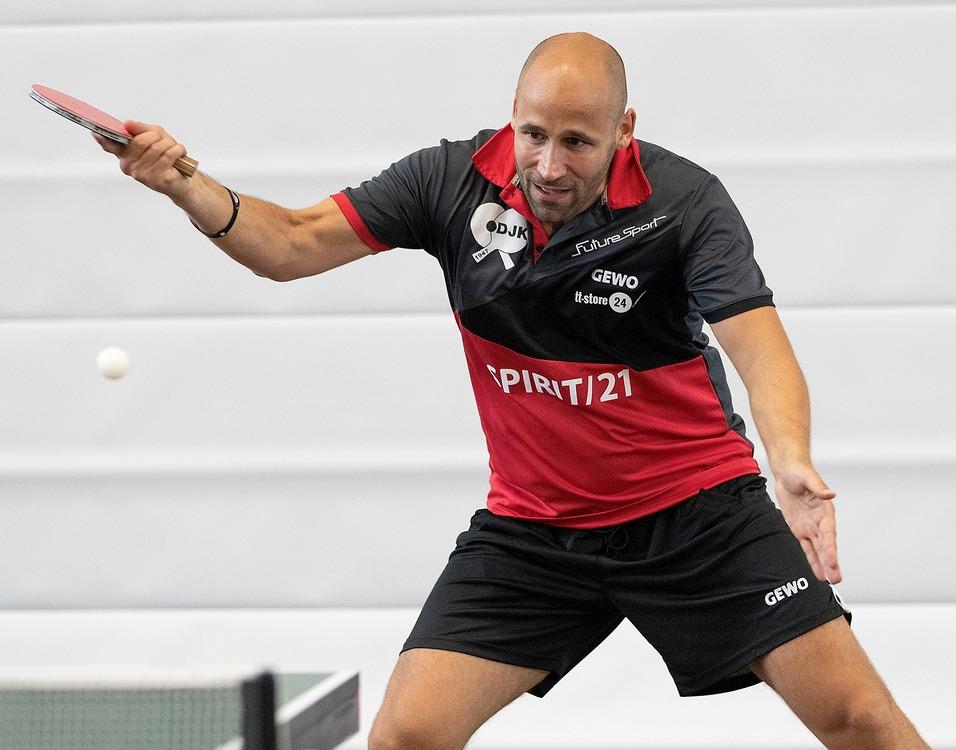 Ein Tischtennisspieler schl&auml;gt einen Ball mit einem Schl&auml;ger w&auml;hrend eines Spiels in einer Sporthalle.