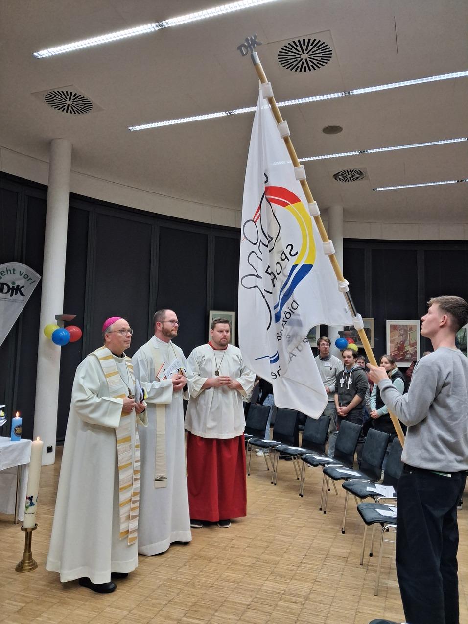 Zwei Geistliche und ein junger Mann halten eine Flagge w&auml;hrend einer Veranstaltung in einem geschlossenen Raum.