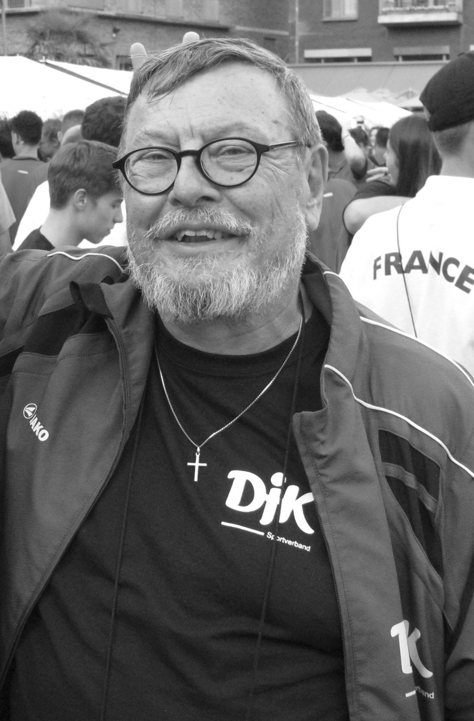 &Auml;lterer Mann mit Brille, Bart und Kreuzkette, tr&auml;gt eine Sportjacke mit dem Schriftzug &bdquo;Dik&ldquo; im Hintergrund.