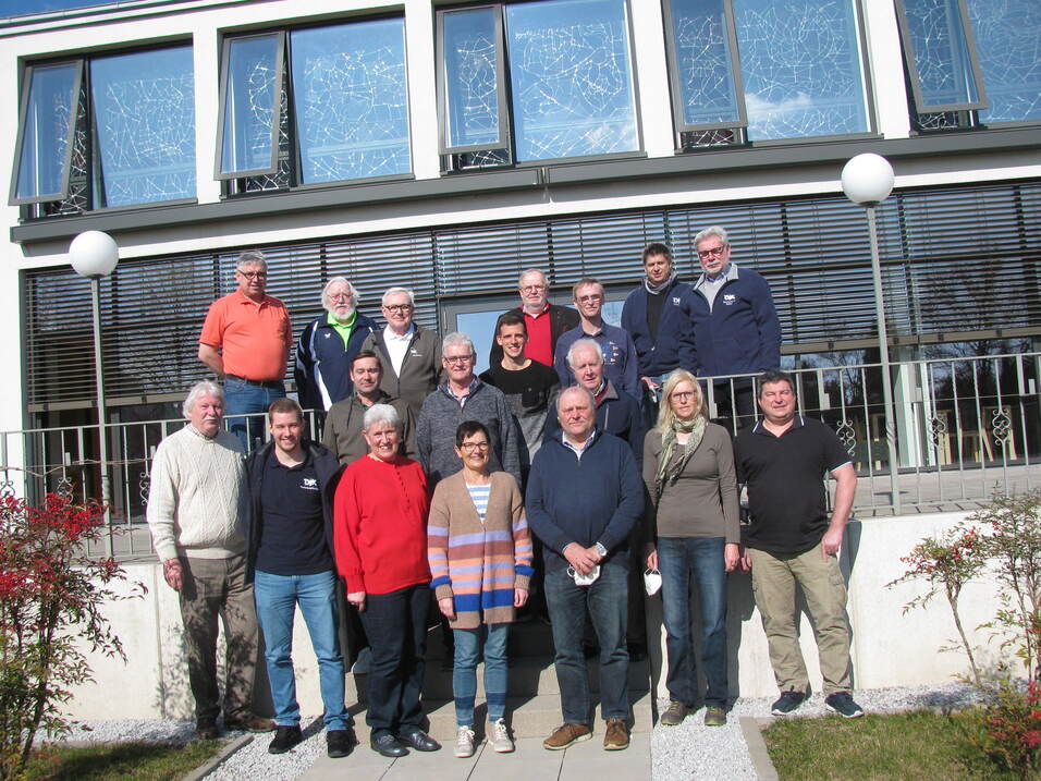 Gruppenfoto von 20 Personen, die vor einem modernen Geb&auml;ude mit Fenstern und Balkon stehen, umgeben von Pflanzen.