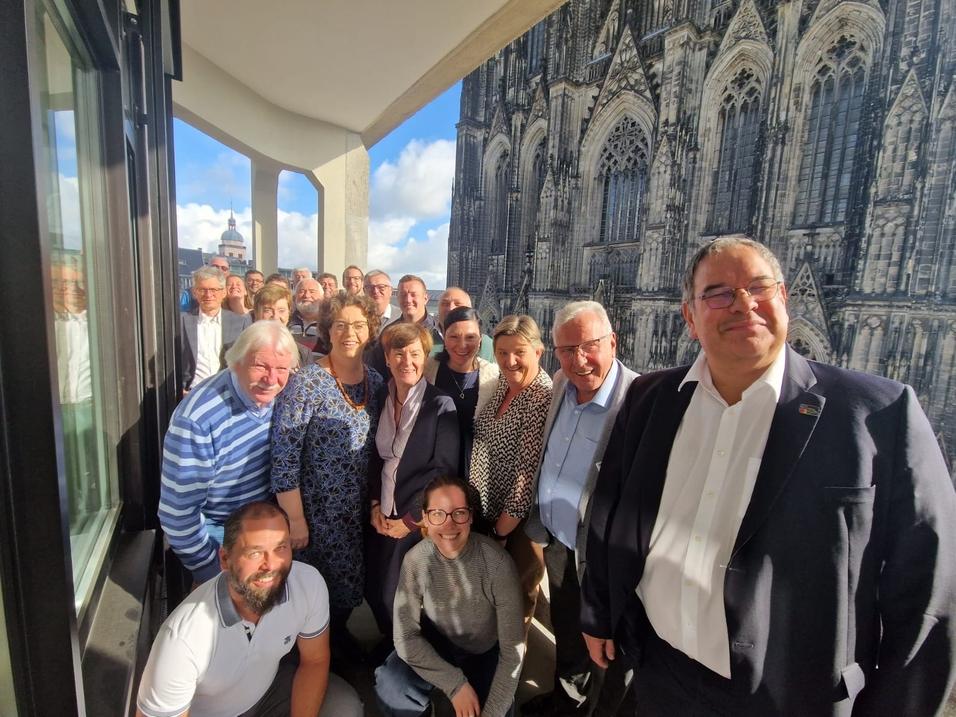 Gruppe von 20 Personen posiert l&auml;chelnd vor einer gro&szlig;en Fensterfront mit Blick auf eine Kathedrale.