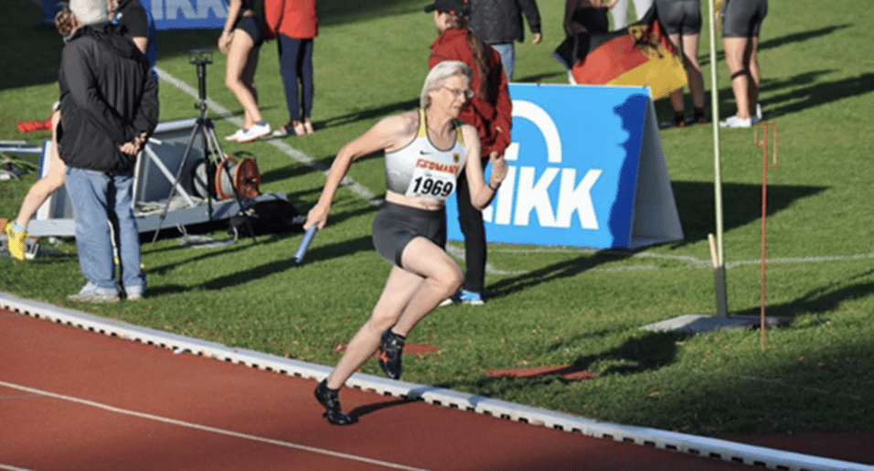 Senioren-Athletin sprintet mit Staffelstab auf der Laufbahn, Zuschauer im Hintergrund, Werbebanner sichtbar.