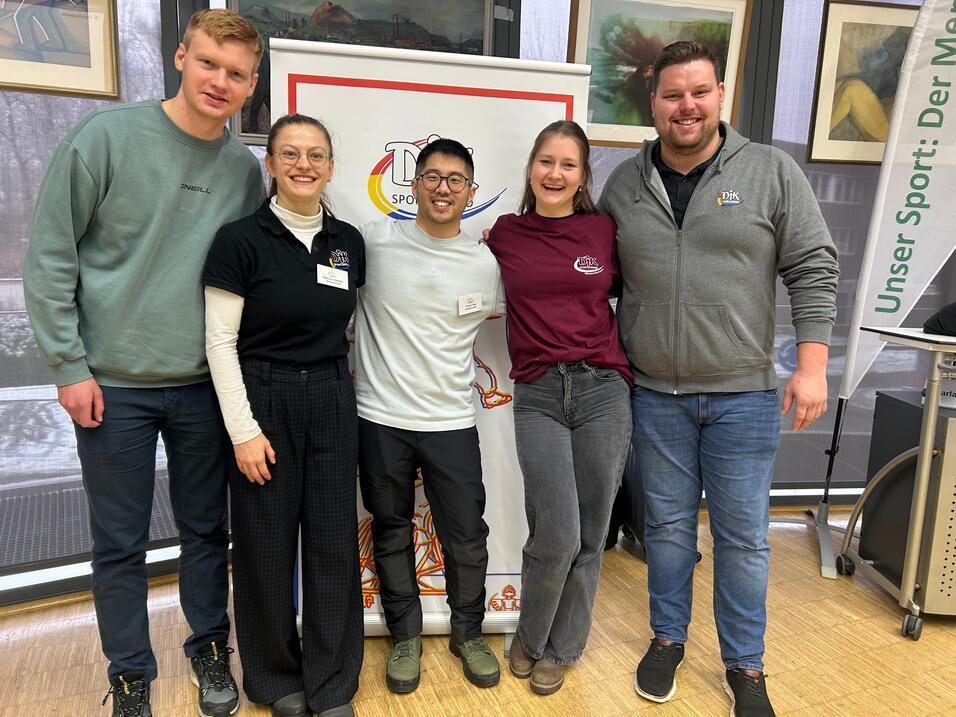 F&uuml;nf Personen stehen l&auml;chelnd vor einem Sportbanner in einem Innenraum. Eine Person tr&auml;gt eine schwarze Bluse, die anderen einheitliche T-Shirts.
