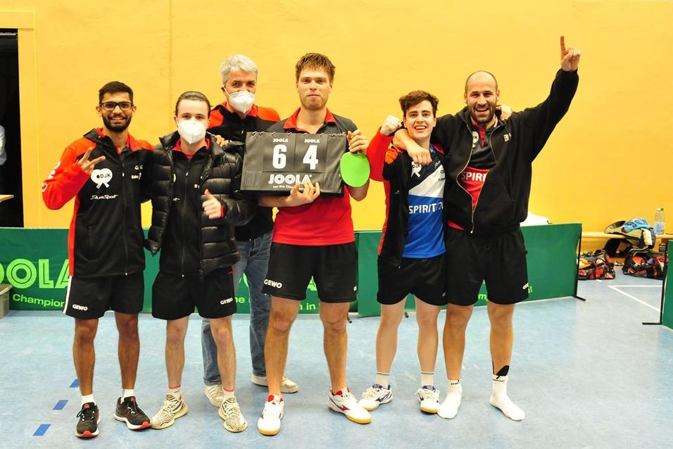 Sechs M&auml;nner stehen l&auml;chelnd zusammen, halten ein Punktetafel mit "6-4" in der Mitte, auf einem Tischtennisplatz.