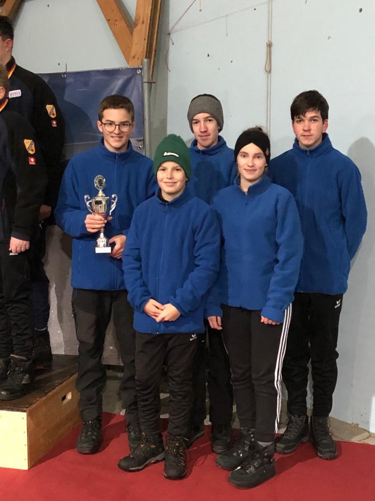 Gruppe von f&uuml;nf Jugendlichen in blauen Jacken, ein Junge h&auml;lt einen Pokal, im Hintergrund eine Sporthalle.