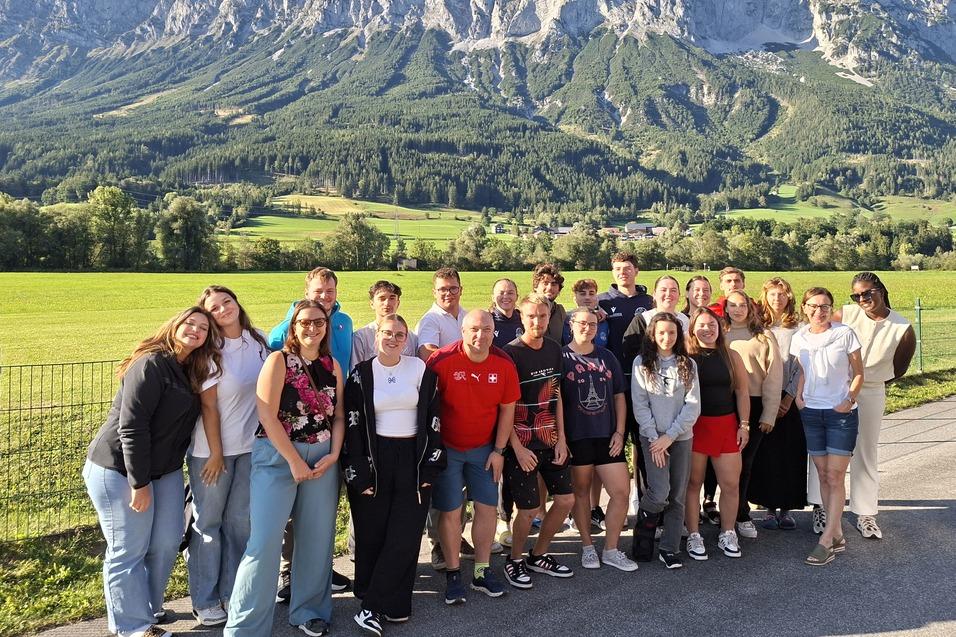 Gruppe von 24 Personen steht vor einer beeindruckenden Berglandschaft an einem sonnigen Tag.