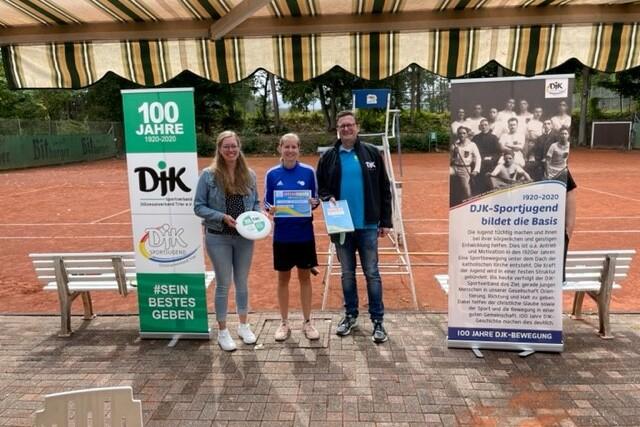 Drei Personen stehen auf einem Tennisplatz, halten Preise und stehen neben Bannern zum 100-j&auml;hrigen Jubil&auml;um der DJK.