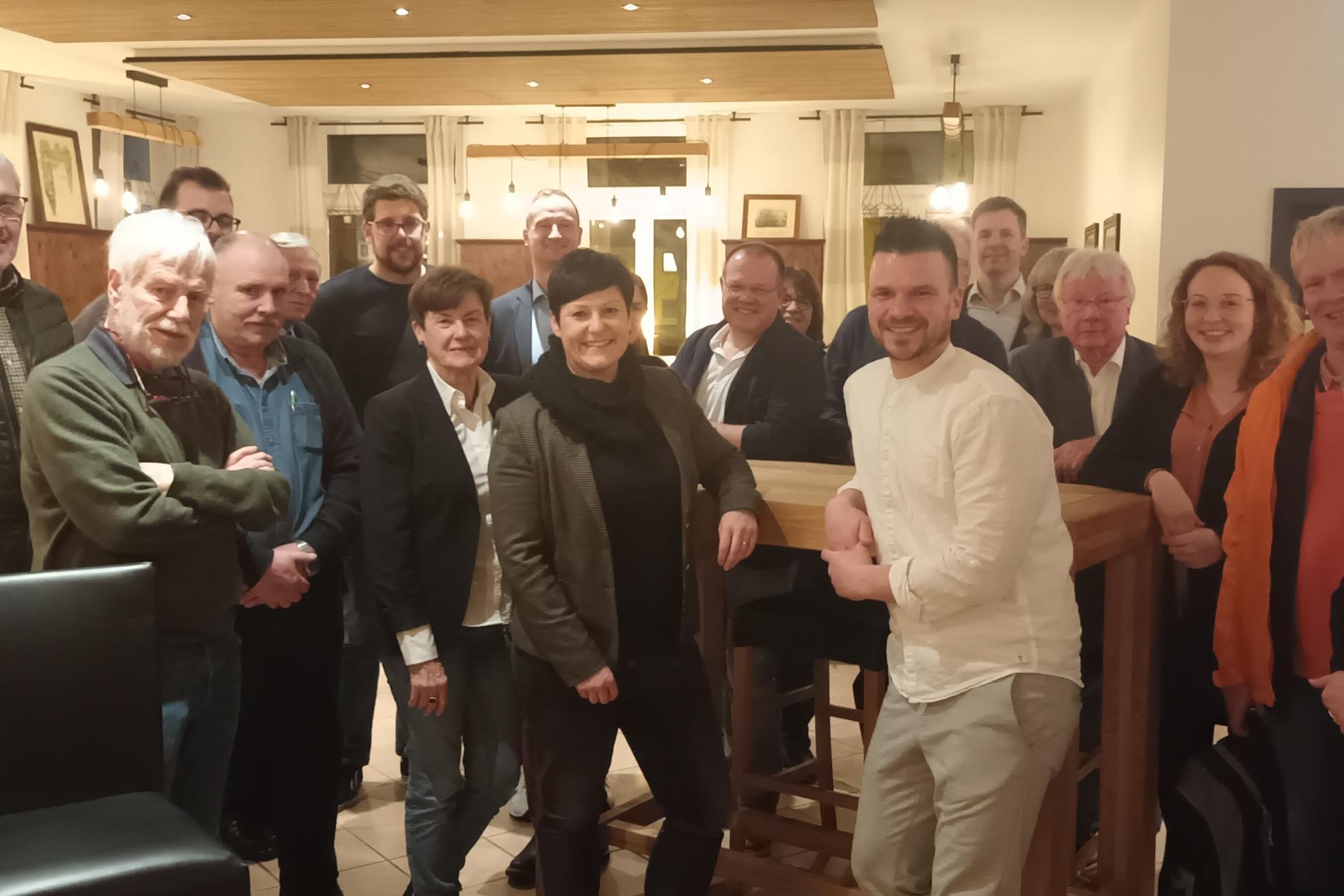 Gruppe von 16 Personen in einem Restaurant, l&auml;chelnd vor einer Bartheke, mit dekorativen Bildern im Hintergrund.