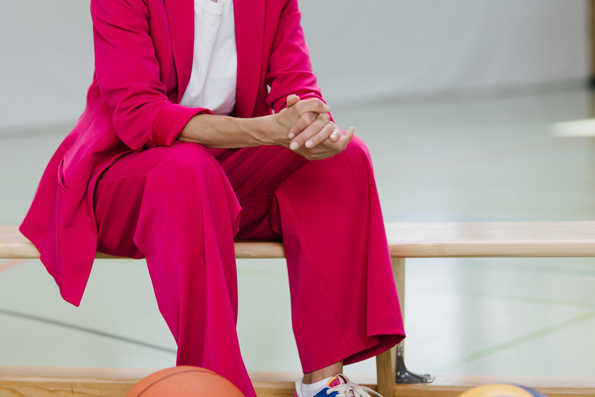 Frau in pinkem Anzug sitzt auf einer Bank, umgeben von Sportb&auml;llen: Basketball, Fu&szlig;- und Volleyball.