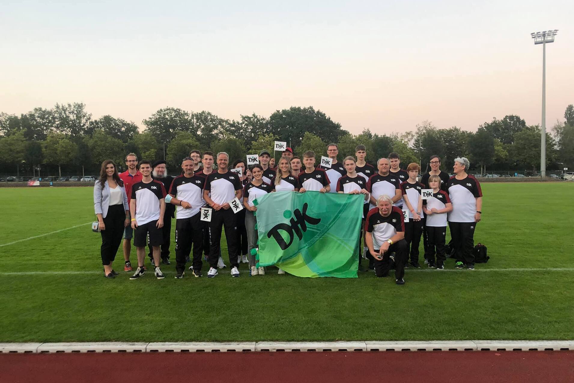 Mannschaft von Sportlern in einheitlicher Kleidung posiert auf einem Rasensportplatz mit einer Flagge und Medaillen.