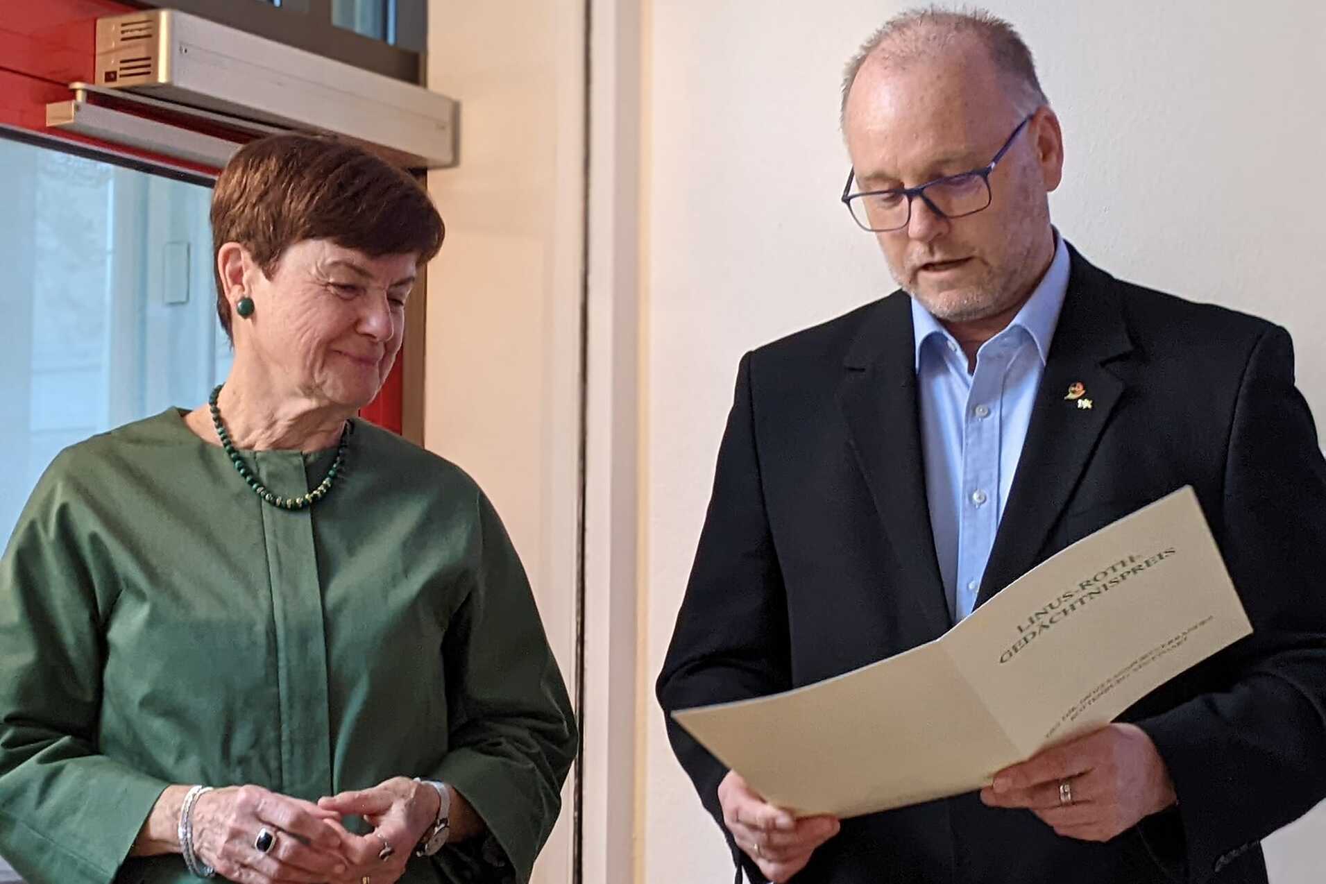 Zwei Personen stehen in einem Raum. Eine Frau in gr&uuml;ner Bluse schaut den Mann an, der ein Dokument liest.