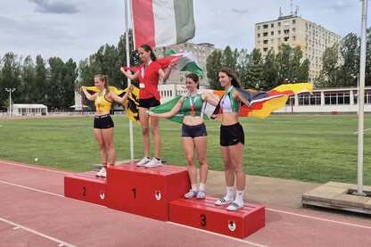 Drei Athletinnen stehen auf dem Siegerpodest, jede mit einer Fahne, im Hintergrund eine Sportanlage und B&auml;ume.