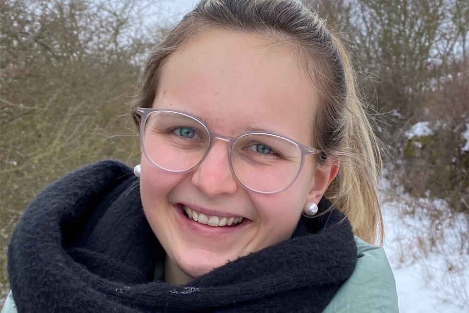 L&auml;chelnde junge Frau mit Brille und Ohrringen, eingepackt in einen schwarzen Schal, beim Spaziergang im Schnee.