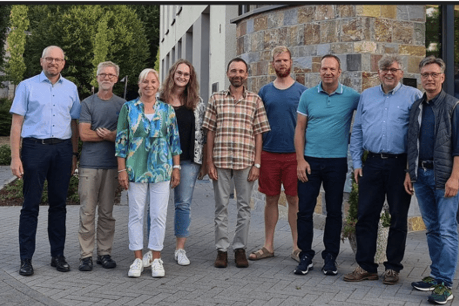 Gruppfoto von neun Personen im Freien, vor einem modernen Geb&auml;ude mit B&auml;umen im Hintergrund.