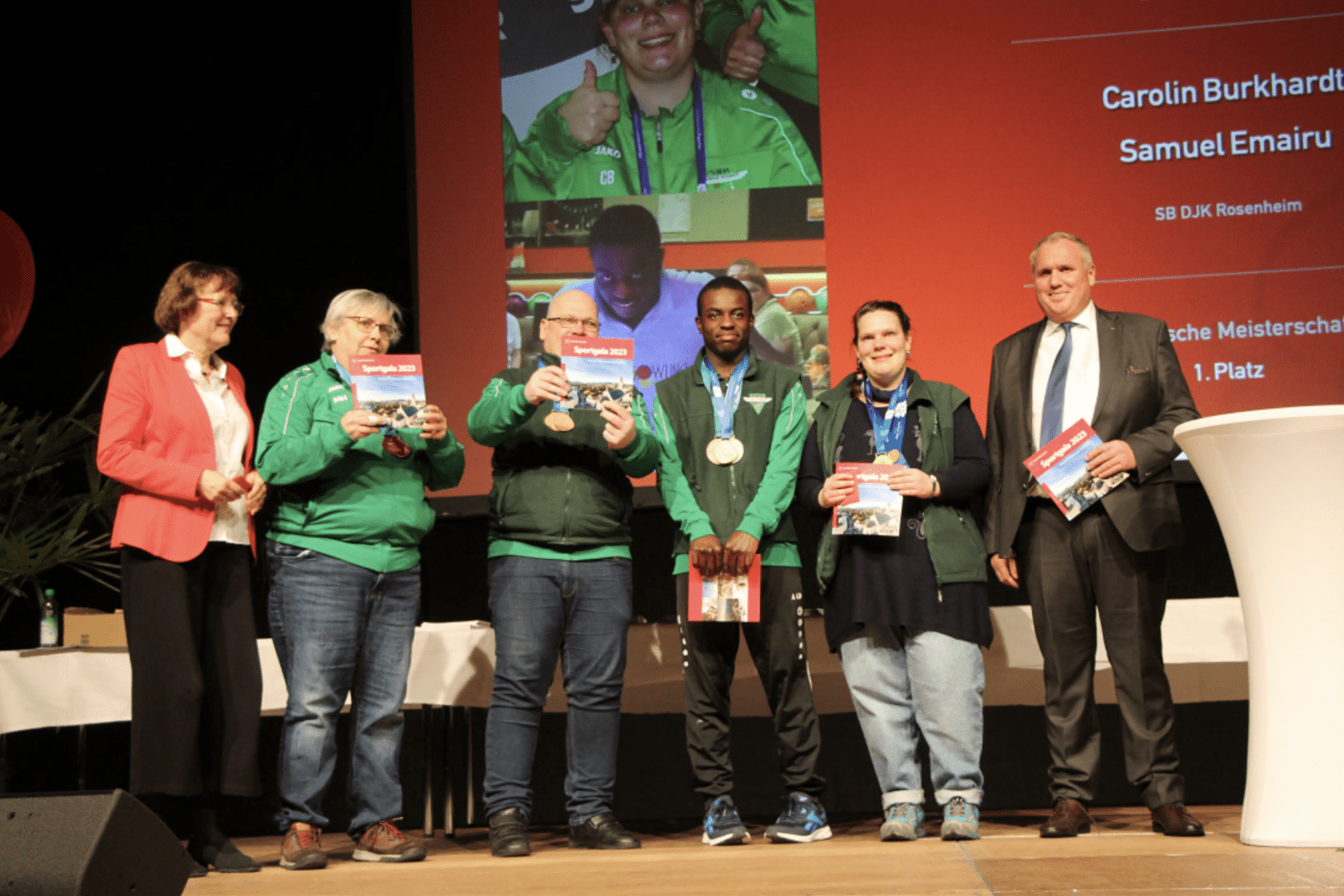 Gruppe von Preistr&auml;gern mit Medaillen und Urkunden auf der B&uuml;hne bei einer Veranstaltung, mit Rednern im Hintergrund.