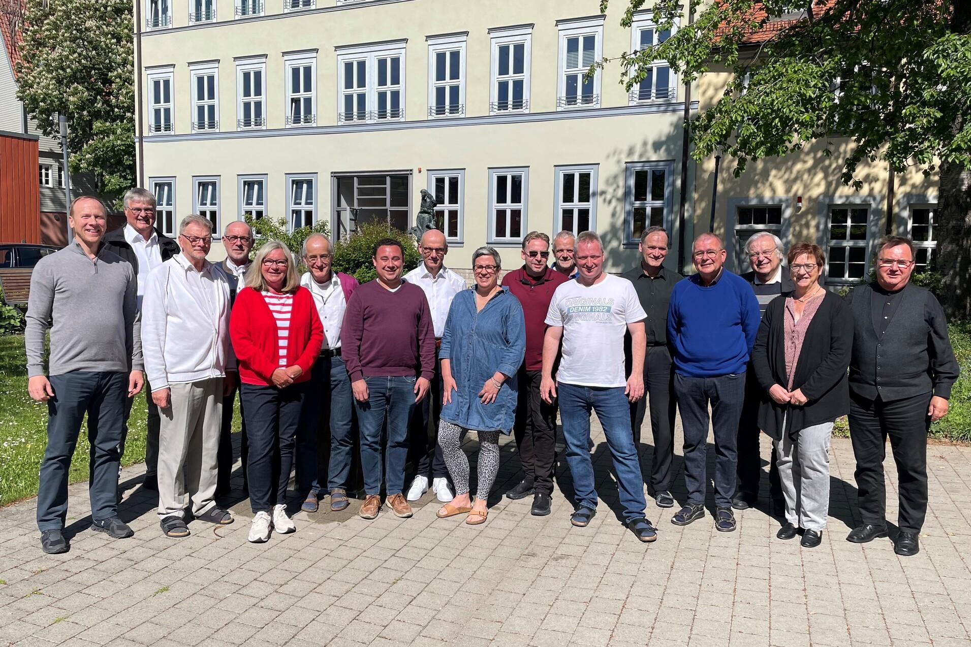 Gruppe von 18 Personen steht auf einem gepflasterten Platz vor einem mehrst&ouml;ckigen Geb&auml;ude mit Fenstern.