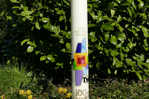 Bunte S&auml;ule mit dem Schriftzug &bdquo;100 Jahre 1920&ldquo; und grafischen Elementen, vor einer gr&uuml;nen Hecke im Freien.