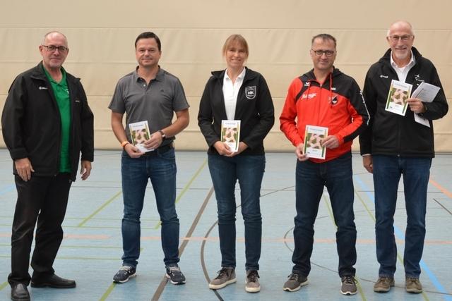 F&uuml;nf Personen stehen in einer Sporthalle, halten Brosch&uuml;ren und l&auml;cheln in die Kamera.