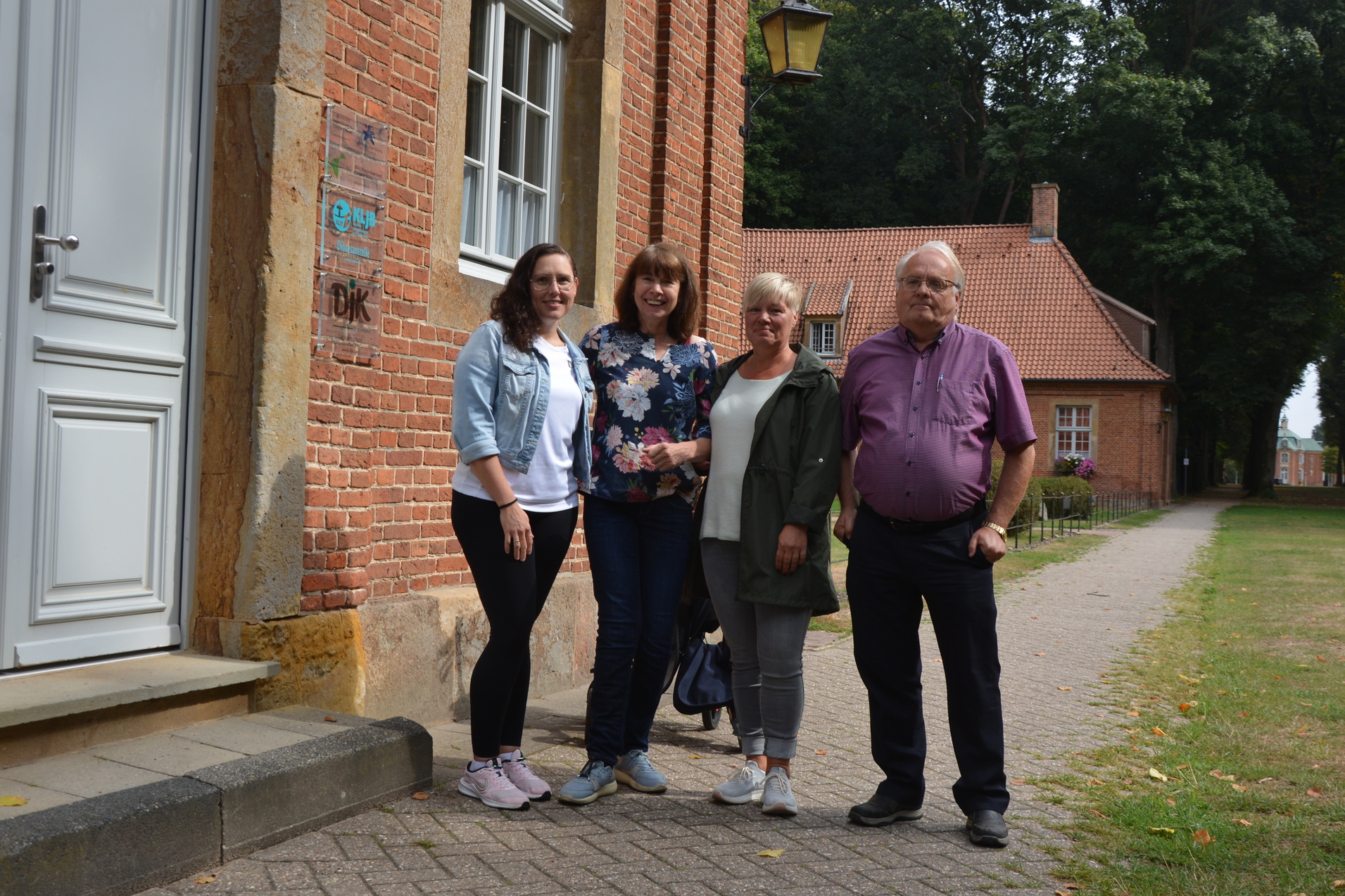 Vier Personen stehen vor einem roten Backsteingeb&auml;ude, einer tr&auml;gt eine hellblaue Jacke. Hintergrund mit Gehweg und B&auml;umen.