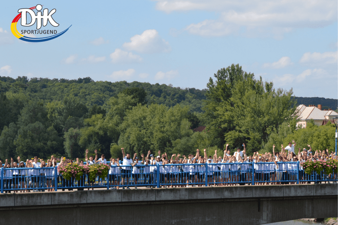Gruppenszene auf einer Br&uuml;cke mit vielen Menschen, die H&auml;nde heben, umgeben von gr&uuml;nen B&auml;umen und blauen Himmel.
