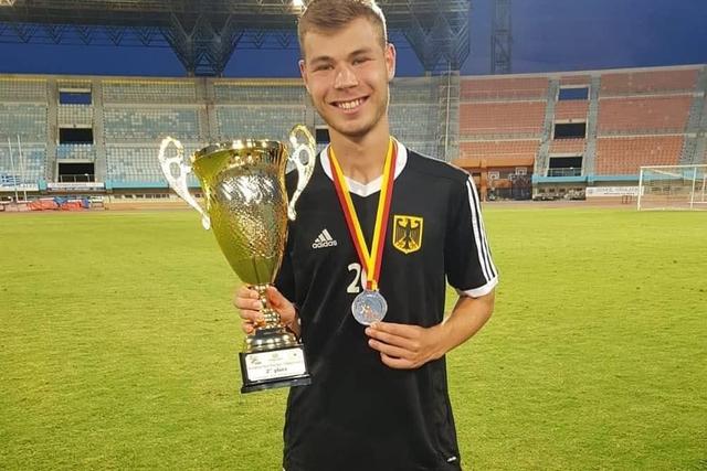 Ein junger Fu&szlig;ballspieler in Schwarz h&auml;lt einen Pokal und eine Medaille auf einem gr&uuml;nen Rasenplatz in einem Stadion.