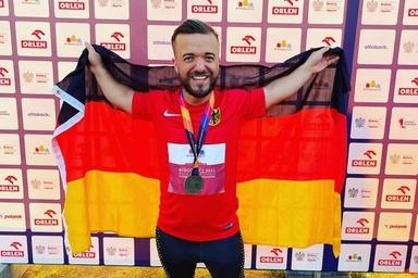 L&auml;chelnder Athlet in rot-schwarzem Trikot h&auml;lt die deutsche Flagge und eine Medaille vor einer Sponsorenwand.