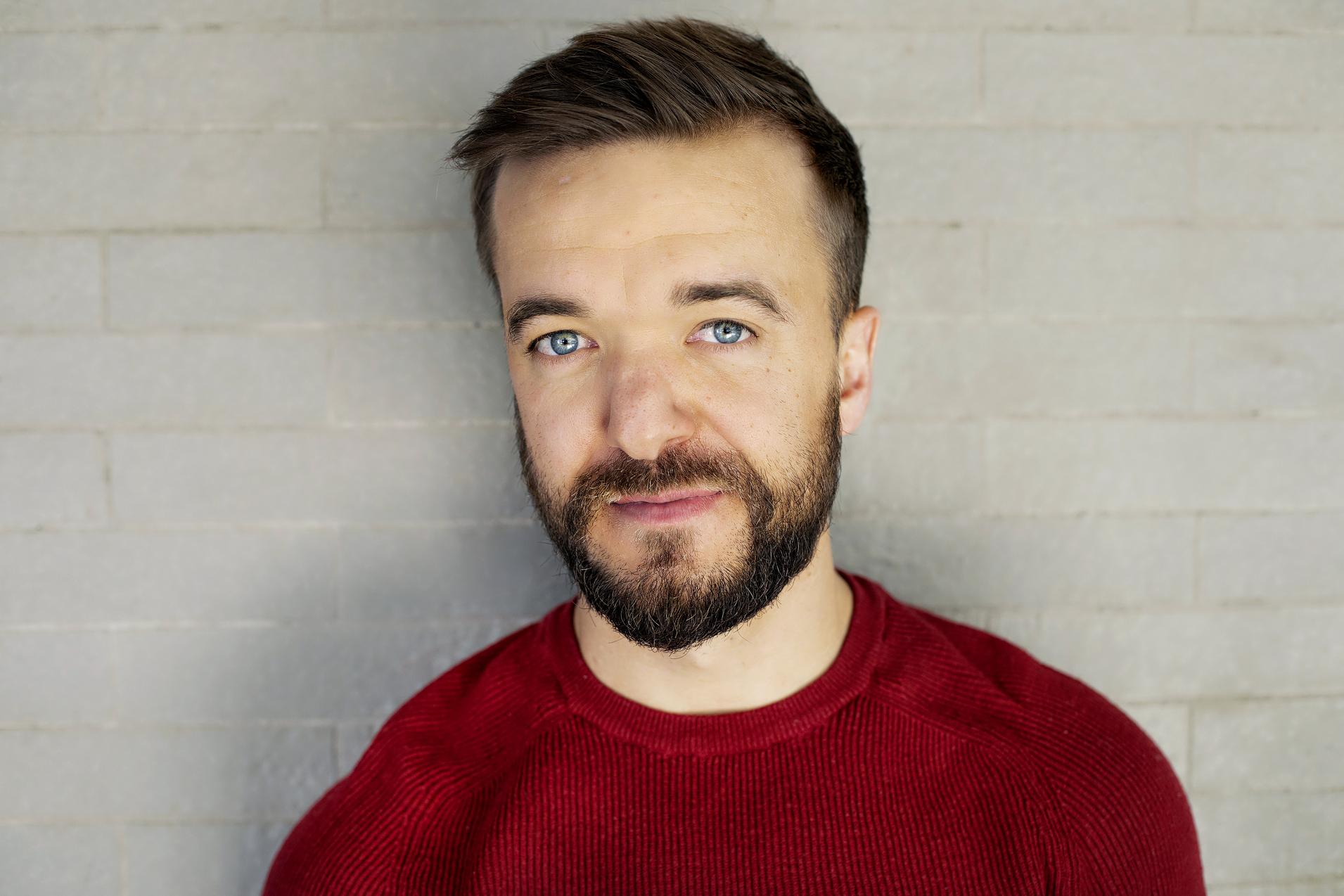 Portr&auml;t eines Mannes mit Kurzhaarfrisur und Bart, l&auml;chelnd vor einer grauen Backsteinwand.