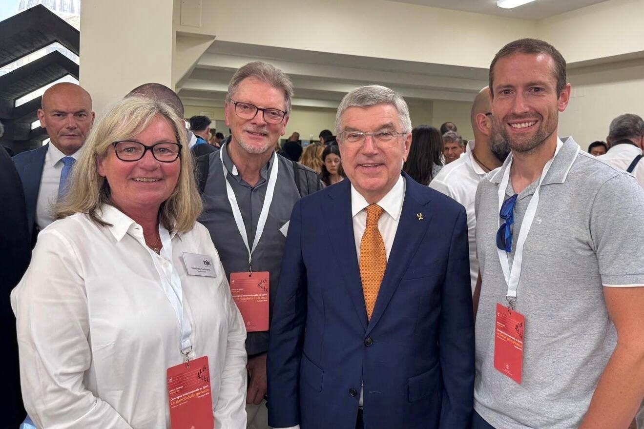 Vier Personen stehen in einem Raum, l&auml;chelnd, mit Namensschildern und einer bekannten Pers&ouml;nlichkeit in Anzug und Krawatte.