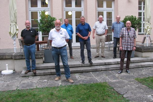Gruppe von sieben M&auml;nnern steht auf einerTerrasse vor einem Geb&auml;ude, umgeben von B&auml;umen und St&uuml;hlen.