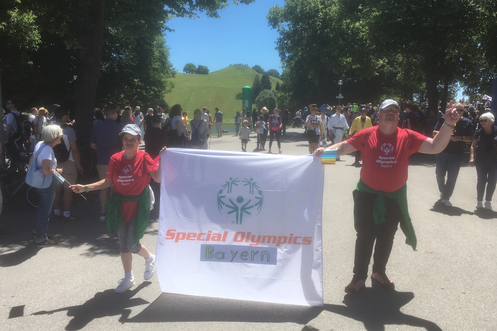 Zwei Menschen in roten T-Shirts halten ein Banner der Special Olympics Bayern in einer belebten Umgebung.