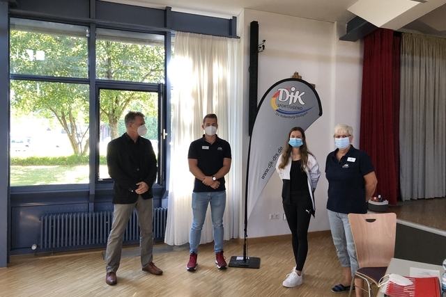 Vier Personen in einem Raum stehen l&auml;chelnd nebeneinander, w&auml;hrend sie Schutzmaske tragen, hinter ihnen ein Banner.