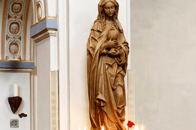 Skulptur der Maria, stehend mit gefalteten H&auml;nden, in einer Kirche, umgeben von Kerzen und einer Rosenbl&uuml;te.