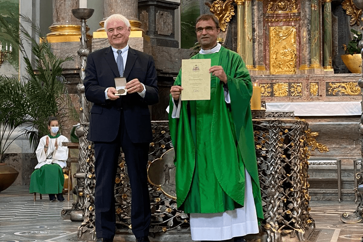 Zwei M&auml;nner stehen in einer Kirche; einer h&auml;lt eine Urkunde, der andere eine Medaille, im Hintergrund eine Person in Kirchenkleidung.